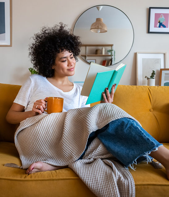 Relaxed, happy African american woman at home living room sitting on the couch, reading a book and drinking tea. 
